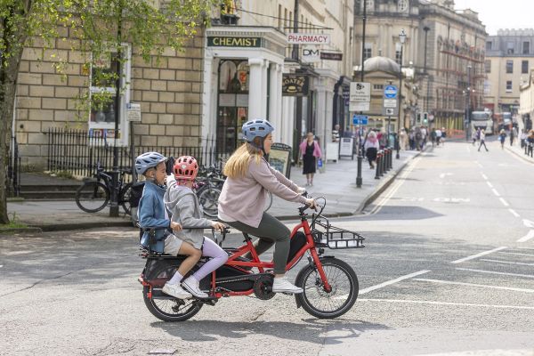 Parent riding a red Tern Quick Haul Long down a city street with two kids on the back