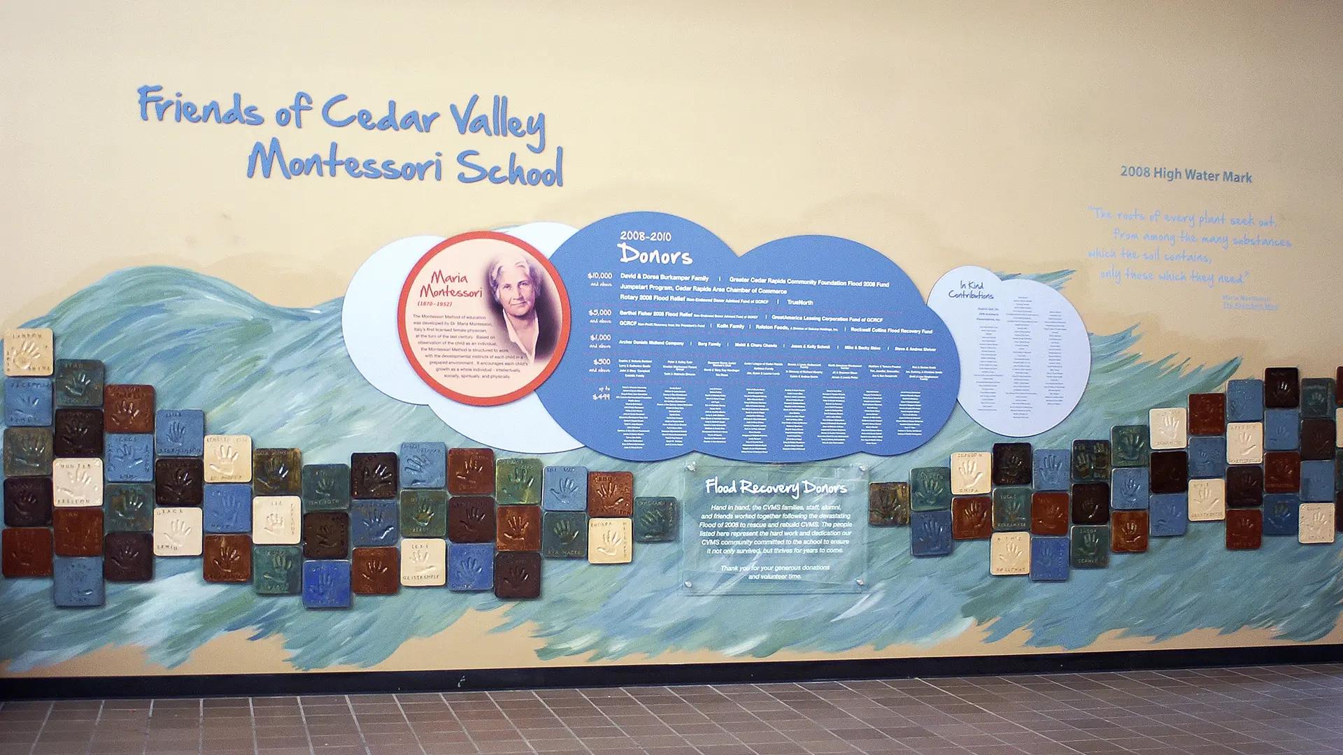School donor recognition wall highlighting flood recovery with a painted river mural and ceramic tiles featuring children’s handprints.