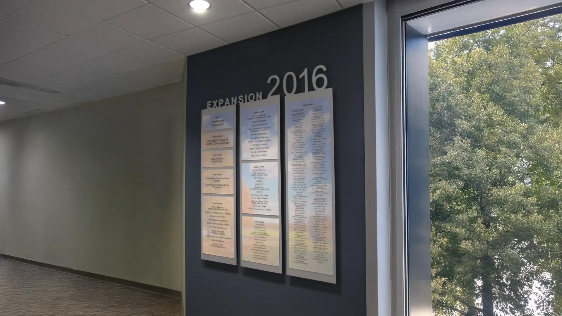 Hospital expansion donor wall displaying 2016 expansion supporters on vertical panels in a light-filled corridor.