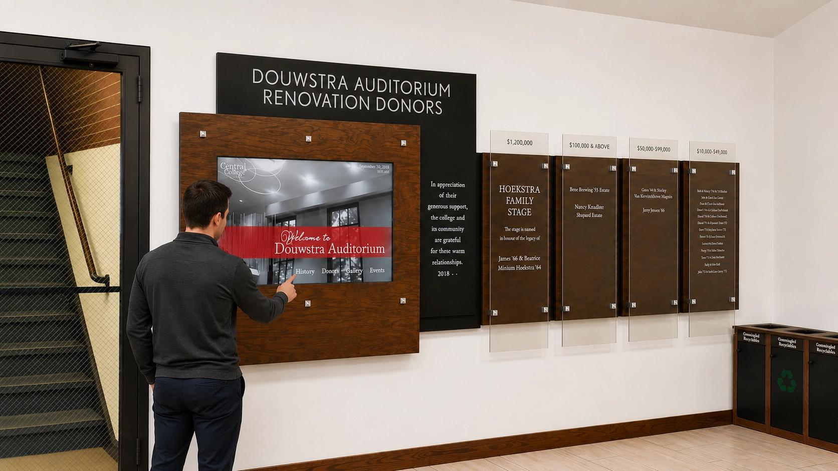 Interactive kiosk donor wall in a college auditorium lobby featuring a touchscreen display framed in wood, surrounded by acrylic donor panels