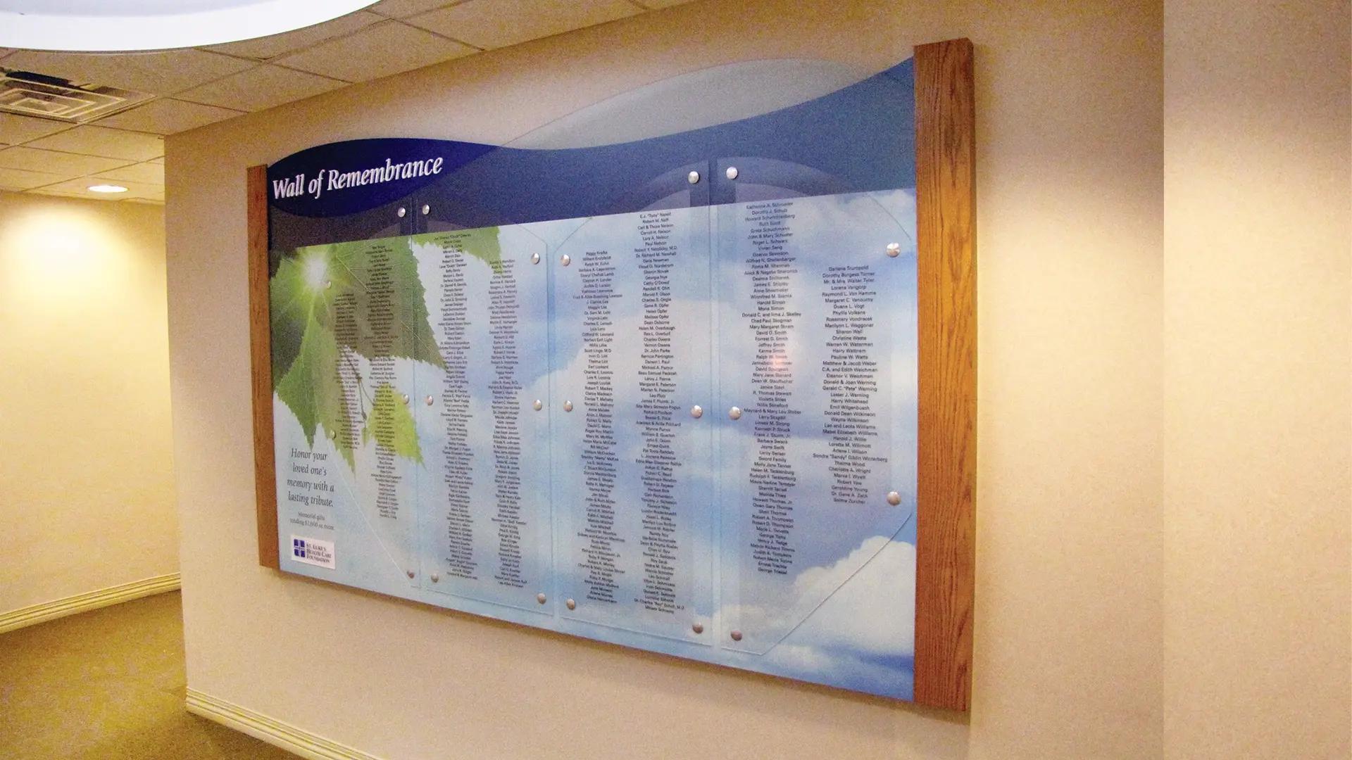 Wall of Remembrance donor wall with printed names on acrylic panels, soft sky imagery, and wood side accents in a quiet corridor.