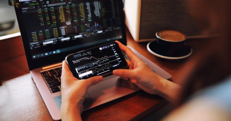 Hands holding a phone with a stock chart app in front of a laptop showing stock quotes, beside a cup of coffee