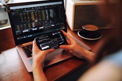 Hands holding a phone with a stock chart app in front of a laptop showing stock quotes, beside a cup of coffee
