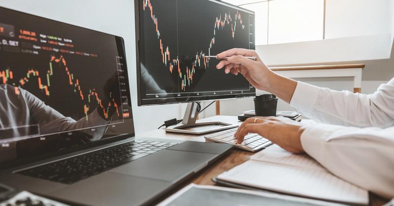 Trader testing a strategy on candlestick charts across laptop and monitor, representing Alpaca paper trading practice