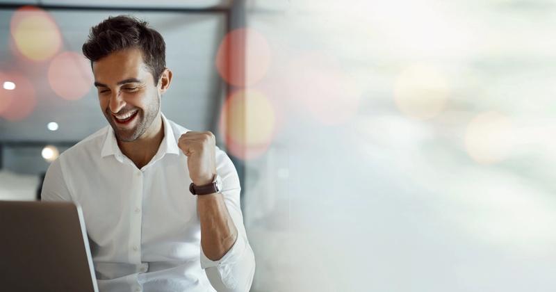 Smiling man in a white shirt making a fist pump while looking at a laptop in a bright office