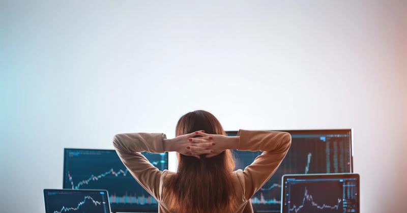 Trader seen from behind with hands behind head, facing two monitors and two laptops showing price charts