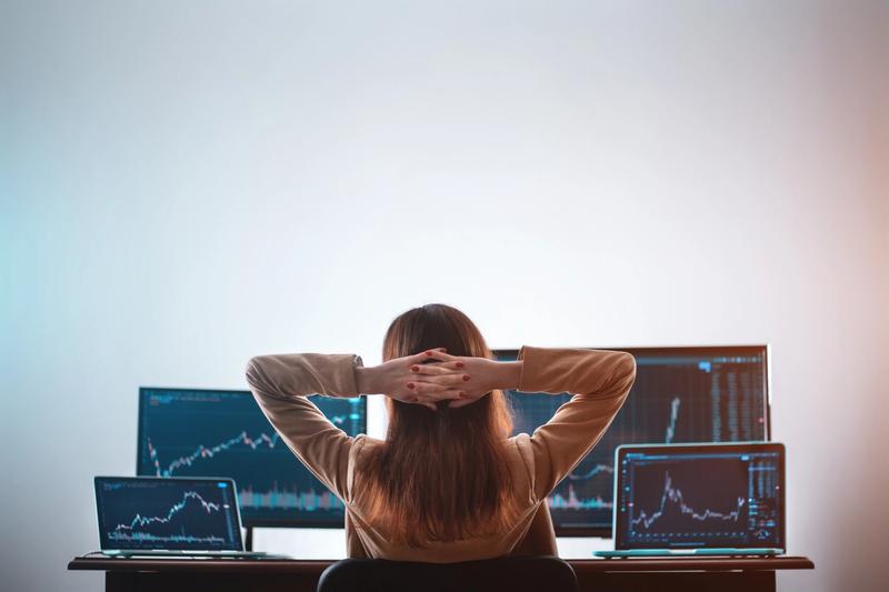 Trader seen from behind with hands behind head, facing two monitors and two laptops showing price charts