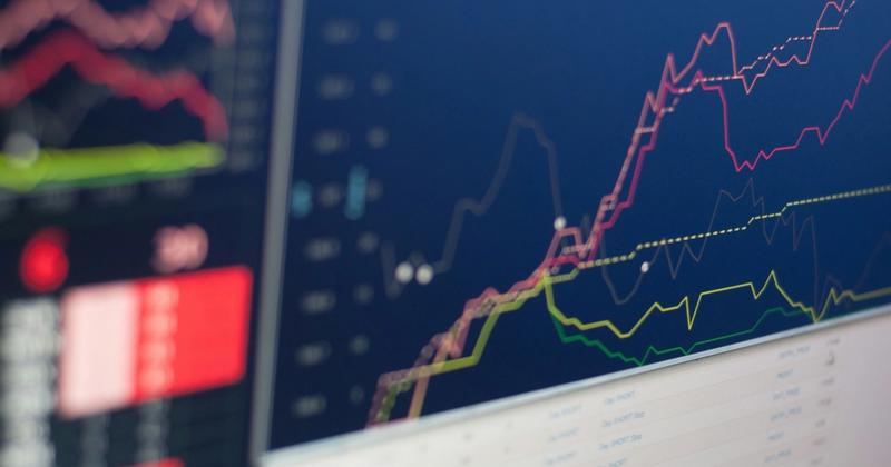 Close-up of a trading monitor showing red, yellow and green line charts on a dark blue screen above an order table