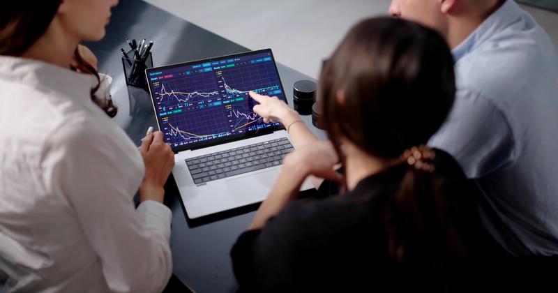 Three people at a desk reviewing a laptop showing four stock chart panels while one points at a price line