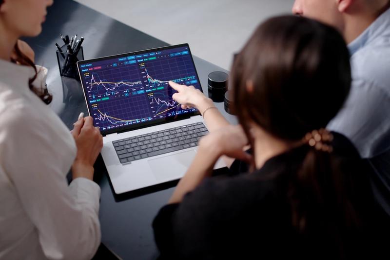 Three people at a desk reviewing a laptop showing four stock chart panels while one points at a price line