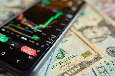 Phone showing a stock chart on top of dollar bills, illustrating Robinhood vs Charles Schwab in 2026
