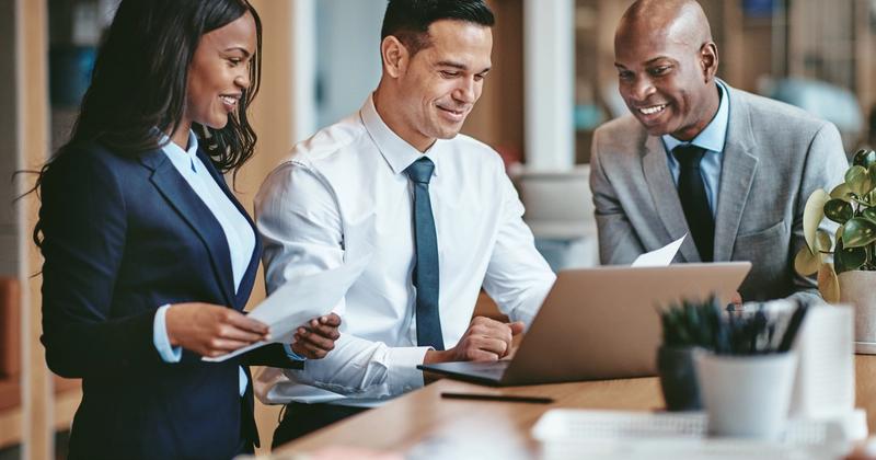 Three colleagues in business attire reviewing papers and a laptop together at an office desk