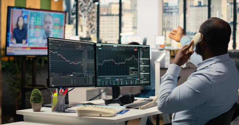 Trader on a desk phone facing two monitors of stock charts, with a TV news broadcast playing across the office