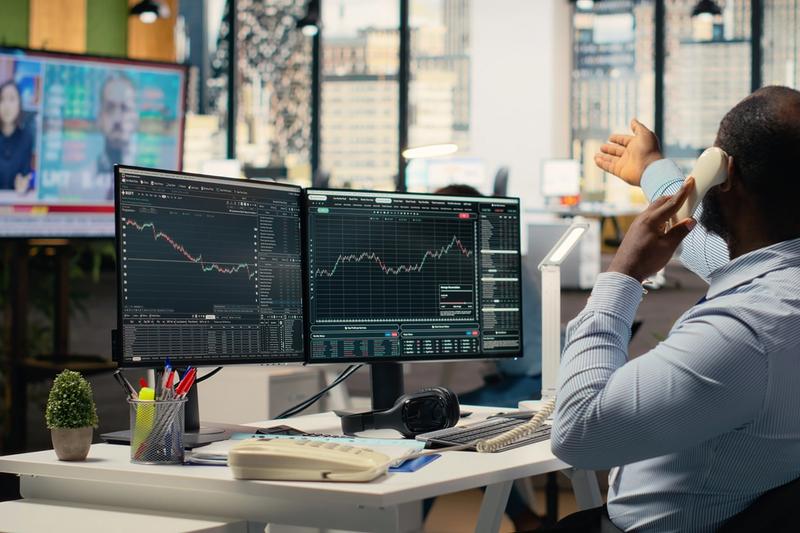 Trader on a desk phone facing two monitors of stock charts, with a TV news broadcast playing across the office