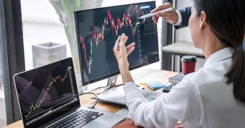 Two people pointing at a candlestick chart on a monitor while a laptop beside it shows past price history