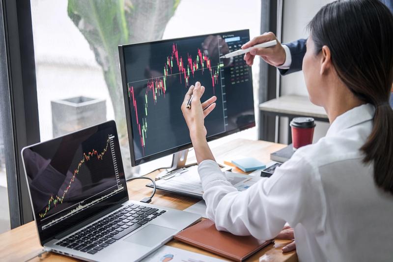 Two people pointing at a candlestick chart on a monitor while a laptop beside it shows past price history