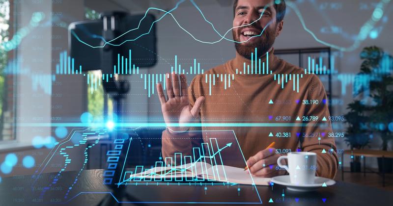 Smiling man at a desk waving to a phone camera, with bar charts and stock price figures overlaid in front of him