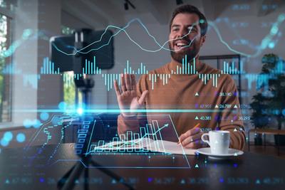 Smiling man at a desk waving to a phone camera, with bar charts and stock price figures overlaid in front of him