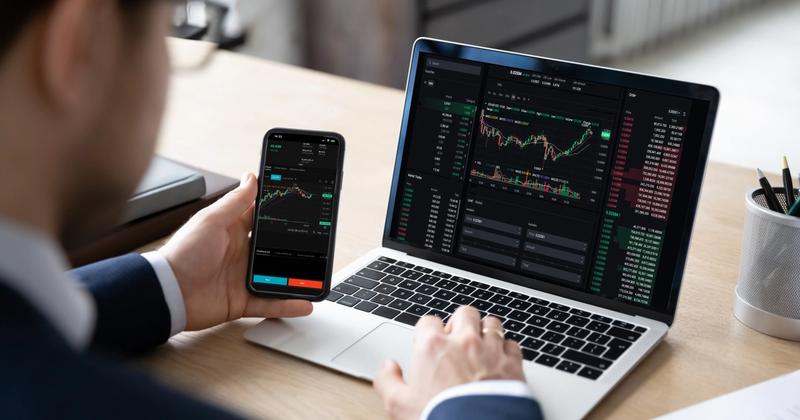 Person in a suit viewing a candlestick trading app on a phone while a laptop shows charts and an order book