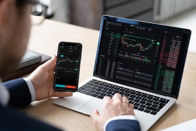 Person in a suit viewing a candlestick trading app on a phone while a laptop shows charts and an order book