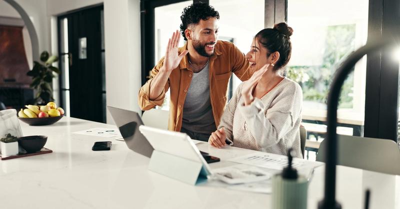 Smiling couple sharing a high five at a kitchen counter with a laptop, tablet, and printed charts