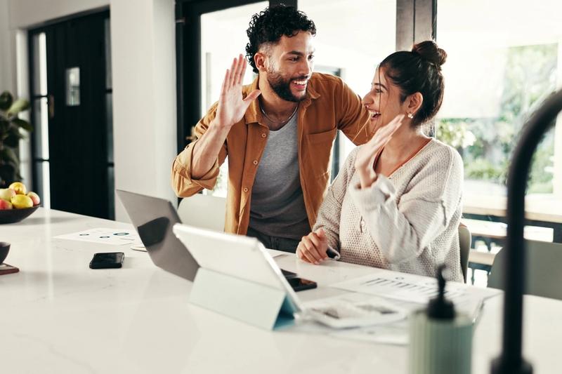 Smiling couple sharing a high five at a kitchen counter with a laptop, tablet, and printed charts