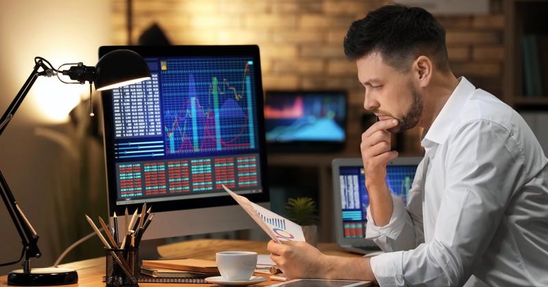 Man at a lamp-lit desk reviewing a printed chart beside a monitor showing stock charts and price tables