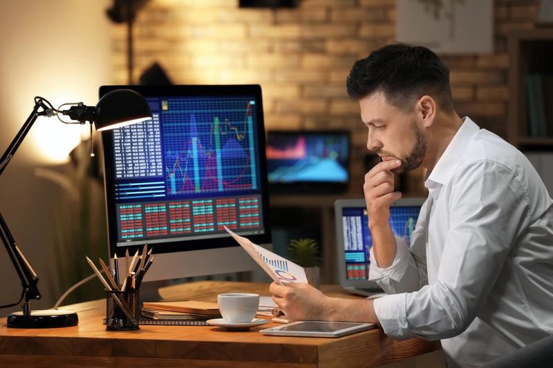 Man at a lamp-lit desk reviewing a printed chart beside a monitor showing stock charts and price tables
