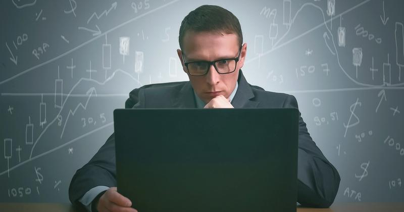 Focused man in glasses and a suit at a laptop, with hand-drawn candlestick charts and numbers behind him