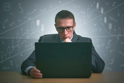 Focused man in glasses and a suit at a laptop, with hand-drawn candlestick charts and numbers behind him