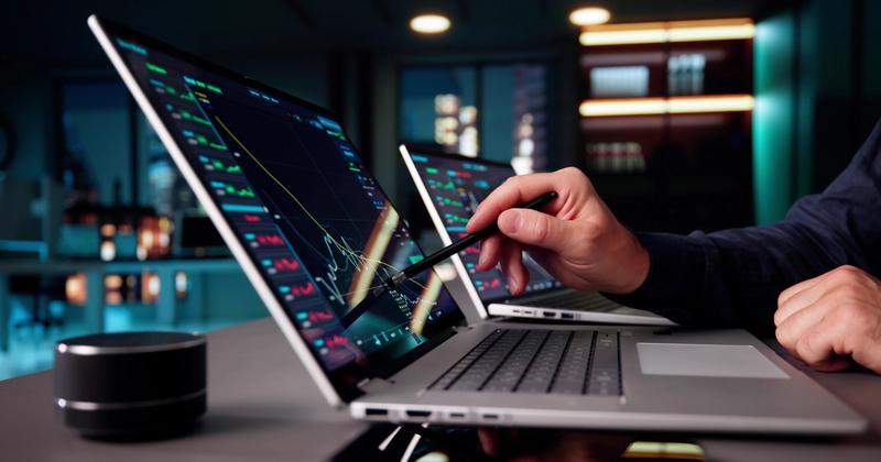 Trader's hand pointing a stylus at stock charts on a laptop screen, with a second laptop behind it in a dim office