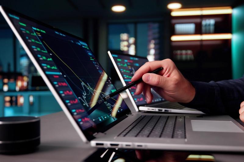 Trader's hand pointing a stylus at stock charts on a laptop screen, with a second laptop behind it in a dim office
