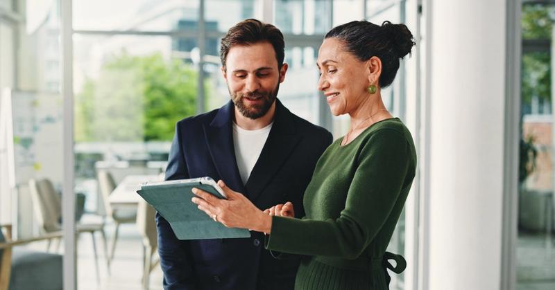 Two smiling colleagues in a bright glass-walled office looking over a tablet together