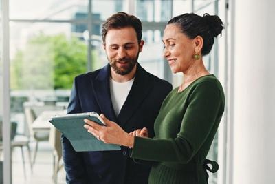 Two smiling colleagues in a bright glass-walled office looking over a tablet together