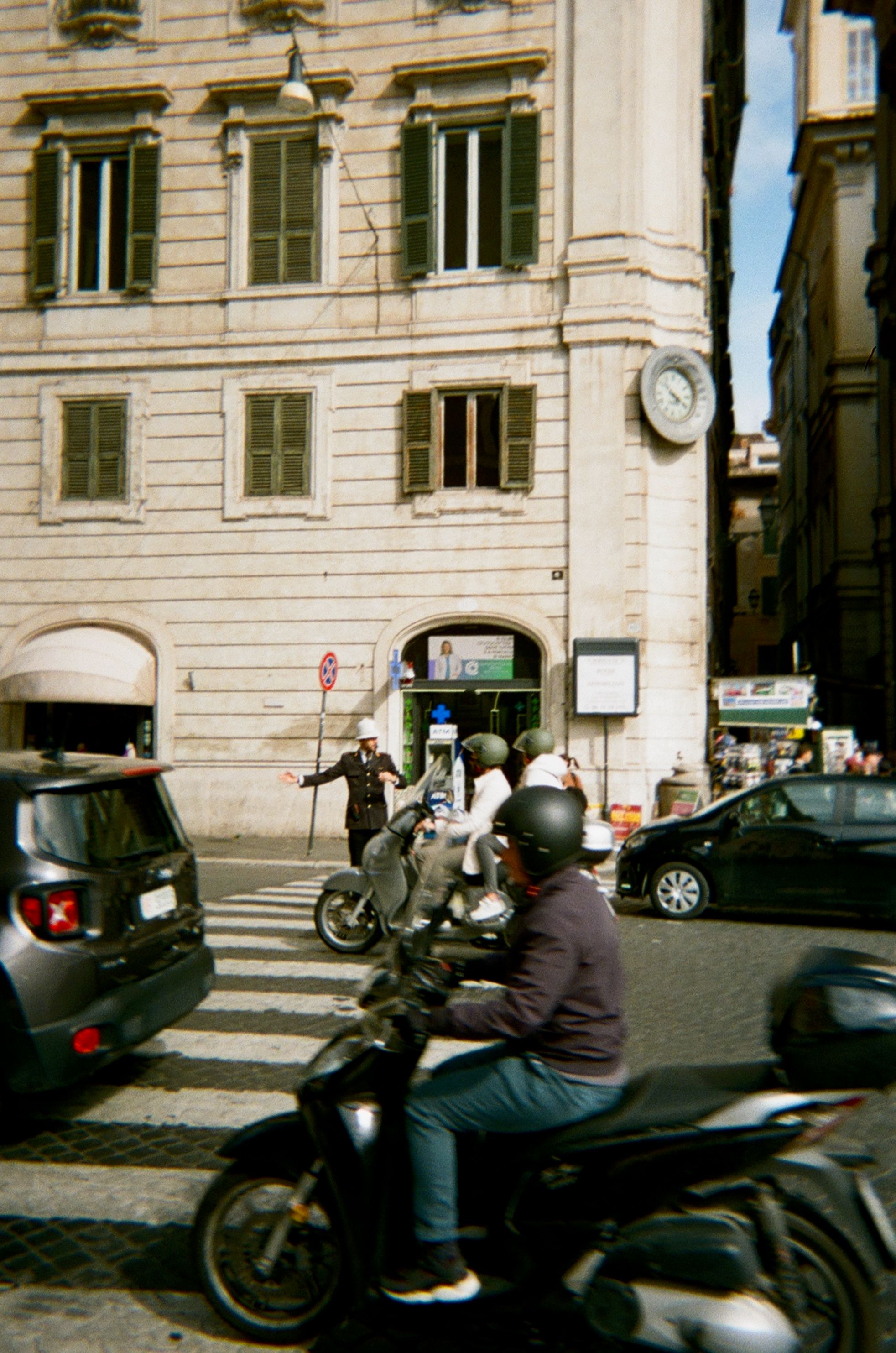 A bustling intersection in Rome, Italy, where a traffic officer directs vehicles and pedestrians. Several motorcyclists and a car are seen moving through the crosswalk. The backdrop features a historic building with green shutters and a large clock mounted on its corner, adding to the city's characteristic charm.