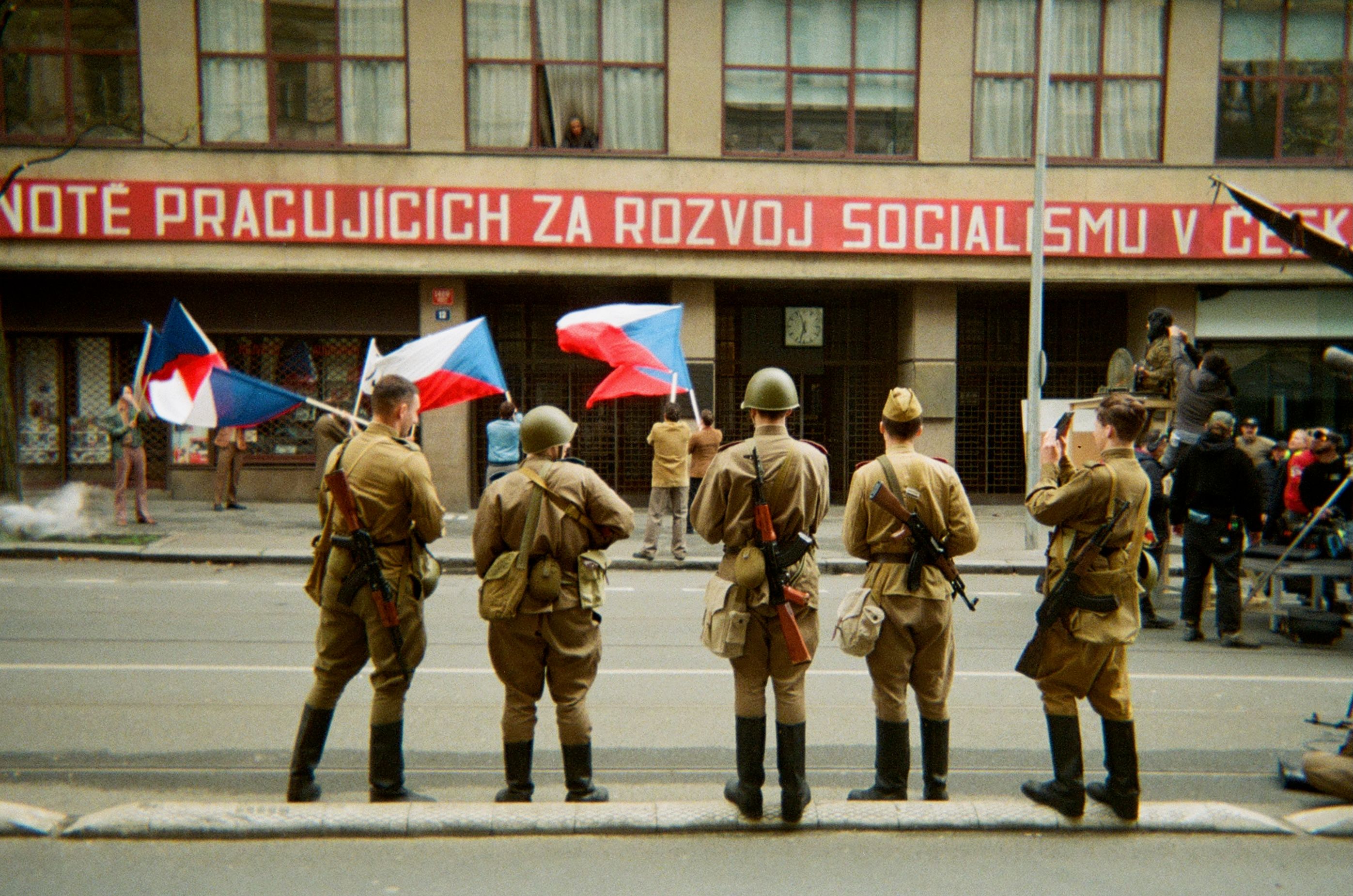 Five soldiers standing in a line with people waving Czech flags in front of them. 
