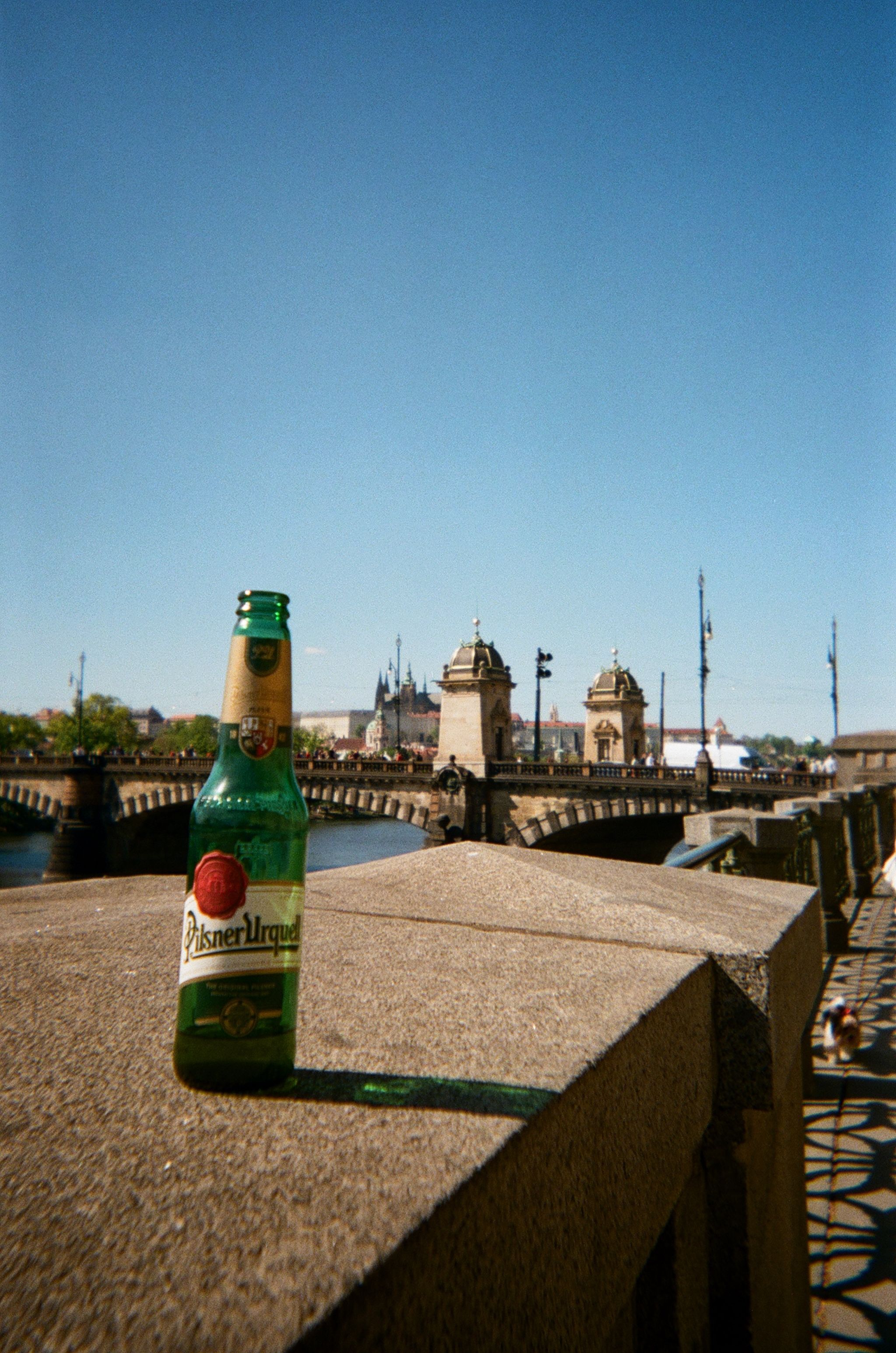 A green bottle of Ilsner Urquell beer sits on a concrete wall overlooking a scenic view.