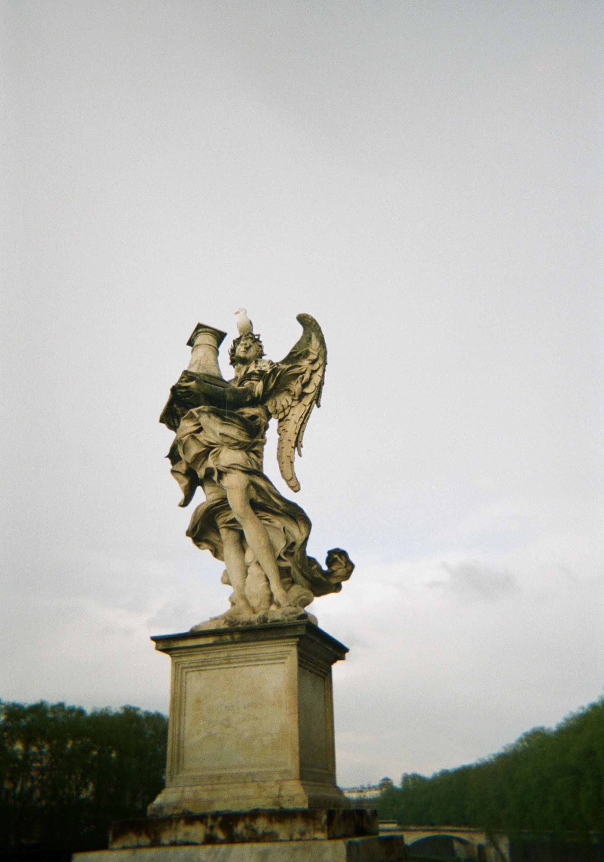 An elegant stone statue of an angel holding a cross stands tall against a pale sky. The angel's wings are spread wide, and a seagull is perched on top of the cross. The statue is set on a sturdy pedestal, with a blurred view of trees and a bridge in the background.