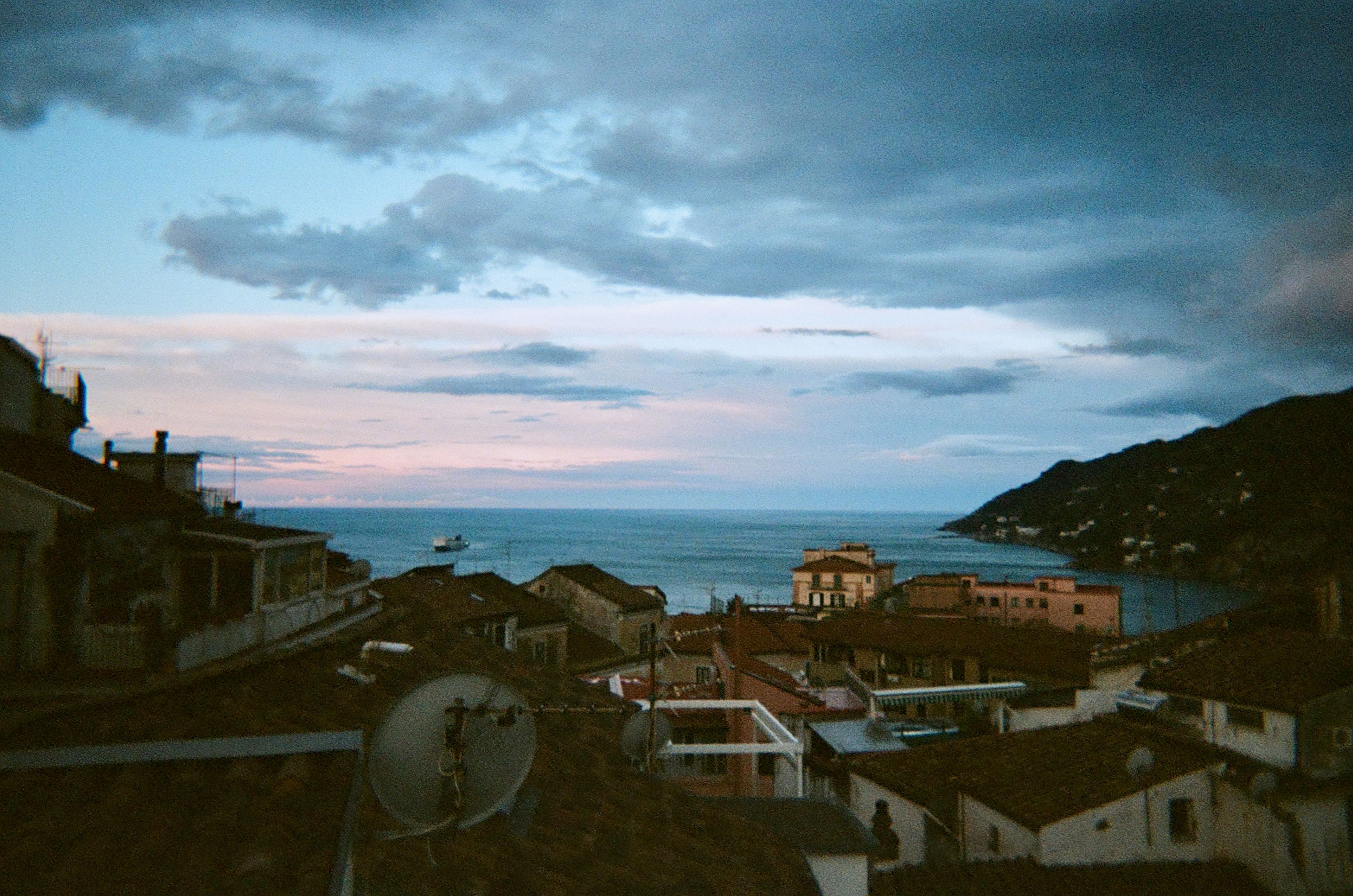 A serene coastal scene at sunrise, featuring the rooftops of a small town overlooking the calm sea. The sky is painted with hues of blue and pink as clouds drift overhead. A boat is visible in the distance on the water, and the silhouette of a mountainous coastline frames the right side of the image