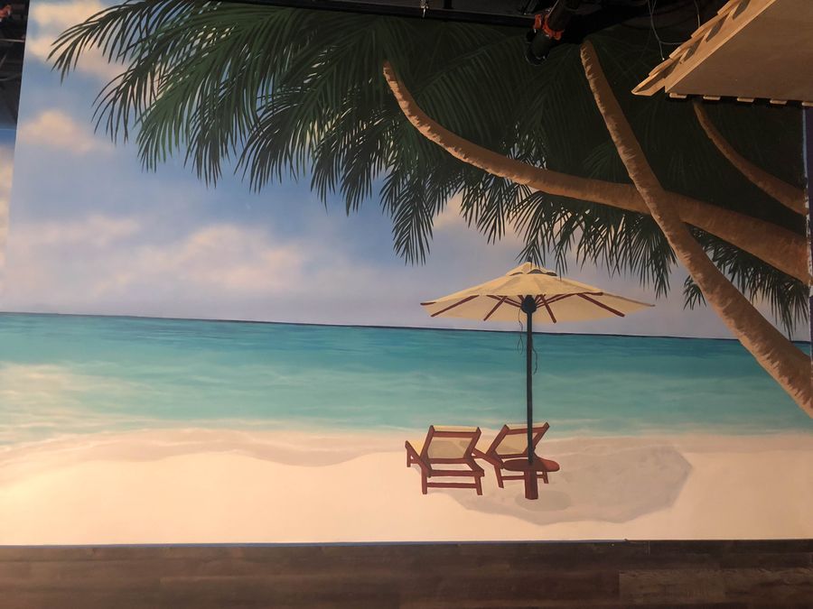 Hand-painted tropical beach wall mural in commercial space with turquoise ocean, lounge chairs, and palm framing.