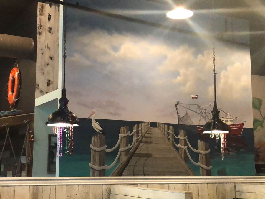 Hand-painted coastal pier mural in Houston restaurant with pelican, Texas shrimp boat, dramatic sky, and rope-post dock.