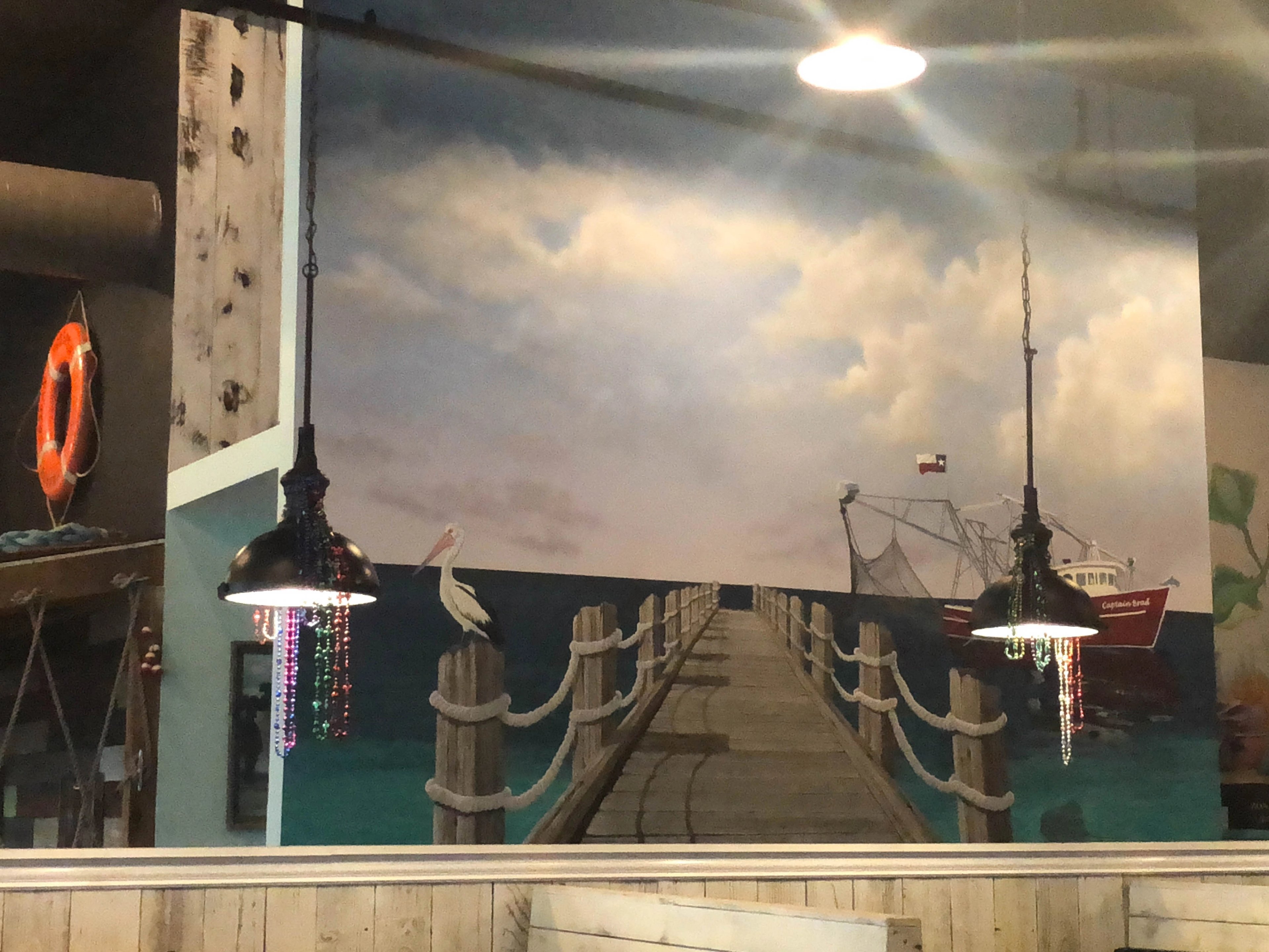 Hand-painted coastal pier mural in Houston restaurant with pelican, Texas shrimp boat, dramatic sky, and rope-post dock.