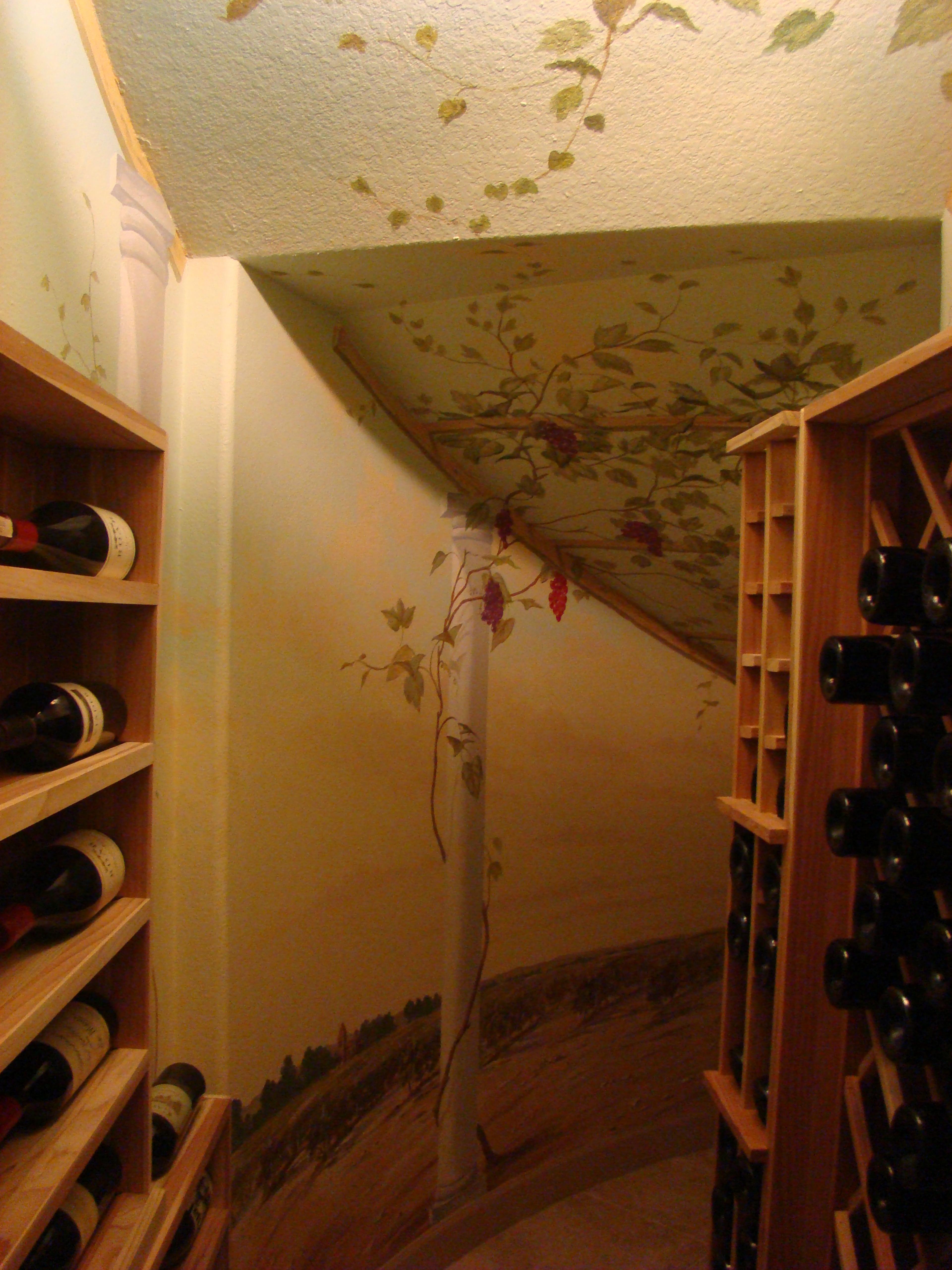 Hand-painted grapevine mural wrapping walls and angled ceiling of residential wine cellar with trompe l'oeil vineyard scene