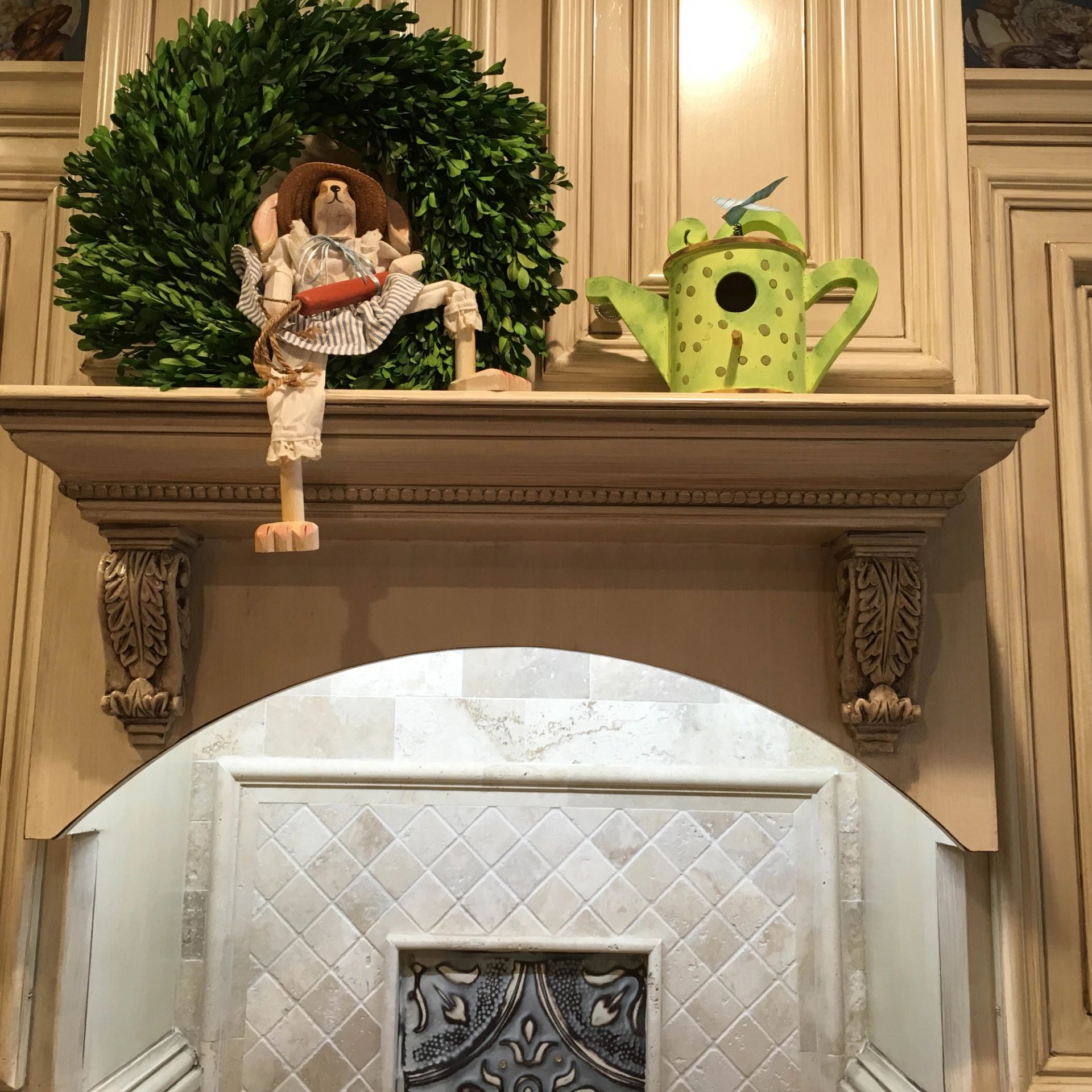 Antiqued glazed cream cabinetry with carved acanthus corbels framing arched travertine backsplash in luxury kitchen.