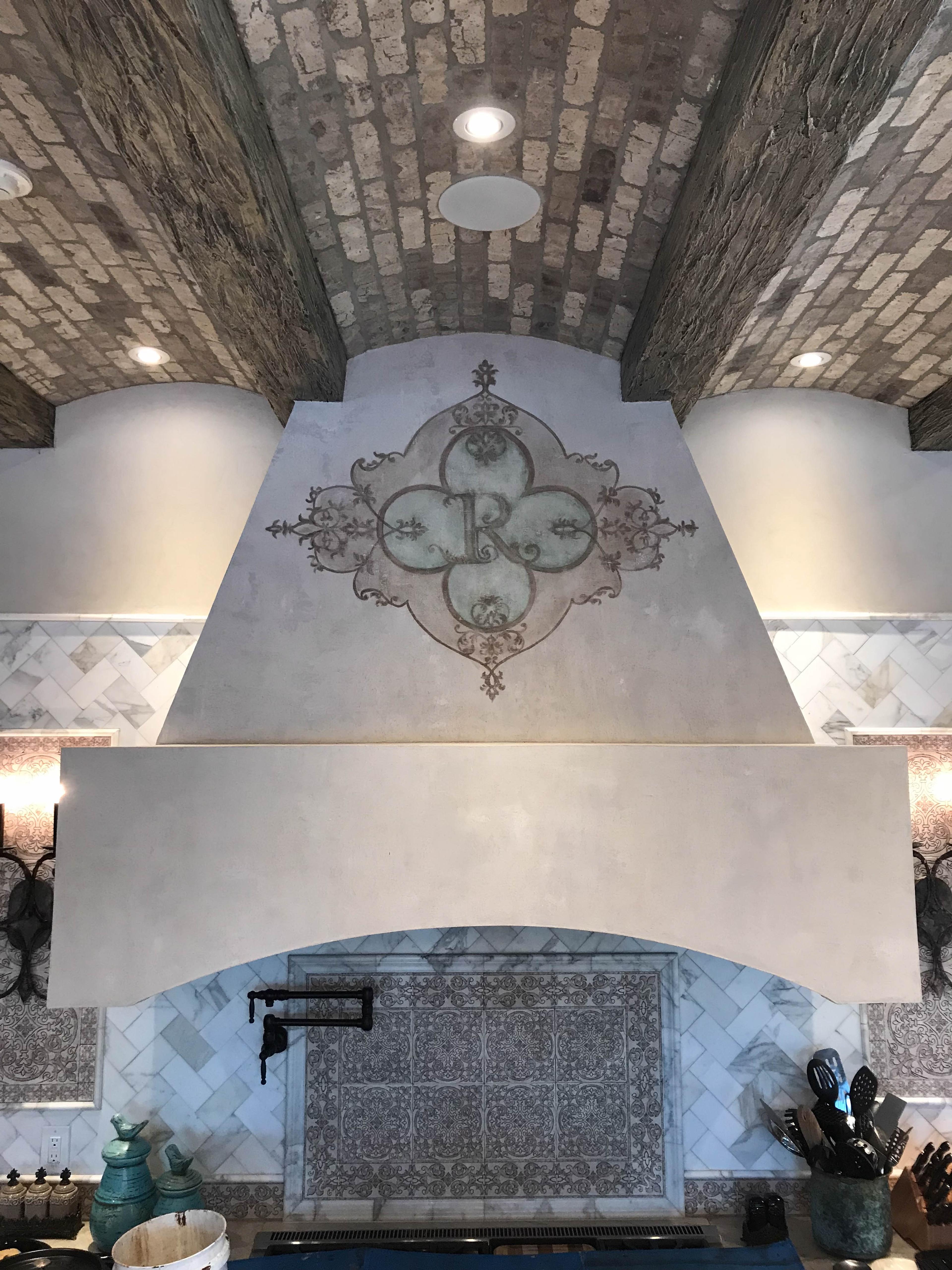 Venetian plaster range hood with hand-painted modello monogram stencil in a grand kitchen with barrel-vault brick ceiling