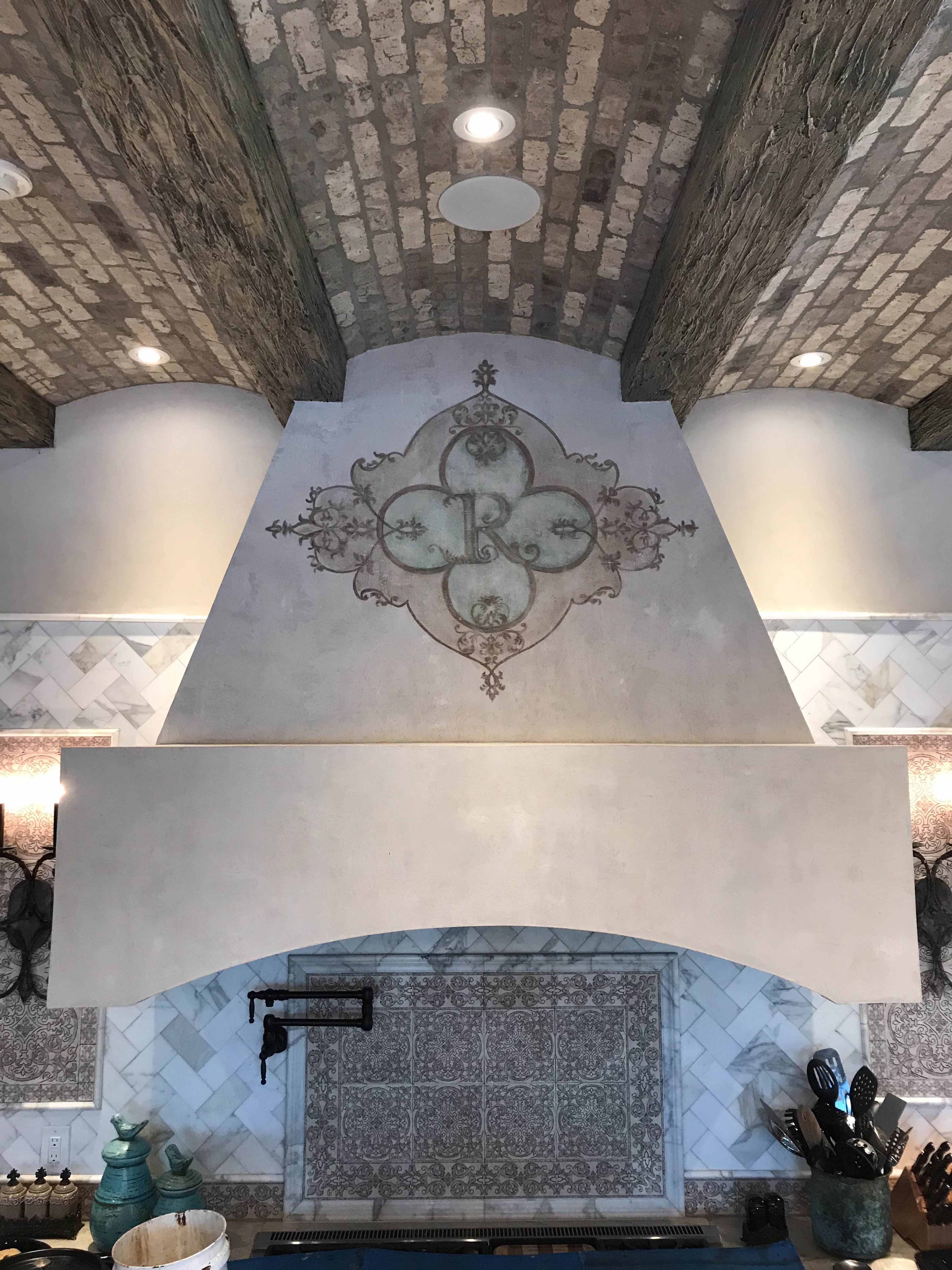 Venetian plaster range hood with hand-painted modello monogram stencil in a grand kitchen with barrel-vault brick ceiling