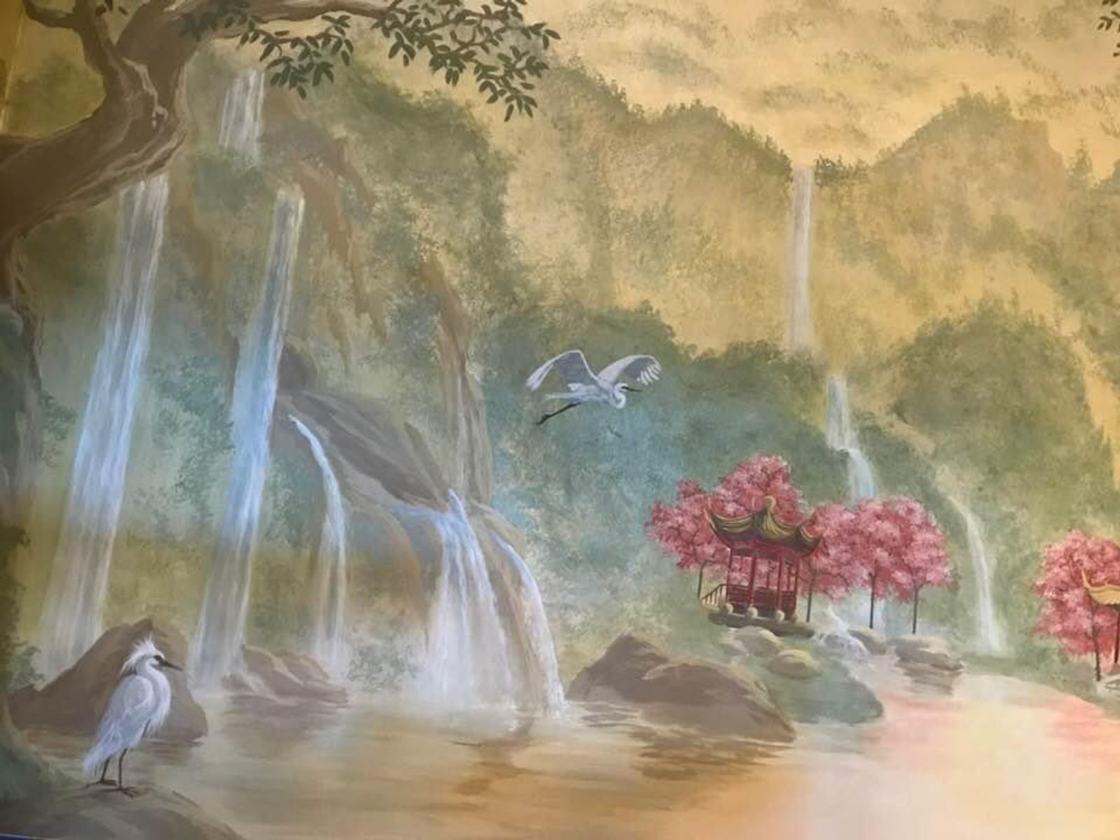 Asian landscape wall mural with hand-painted waterfalls, pagoda, cherry blossoms, and egrets in warm ochre and sage tones.
