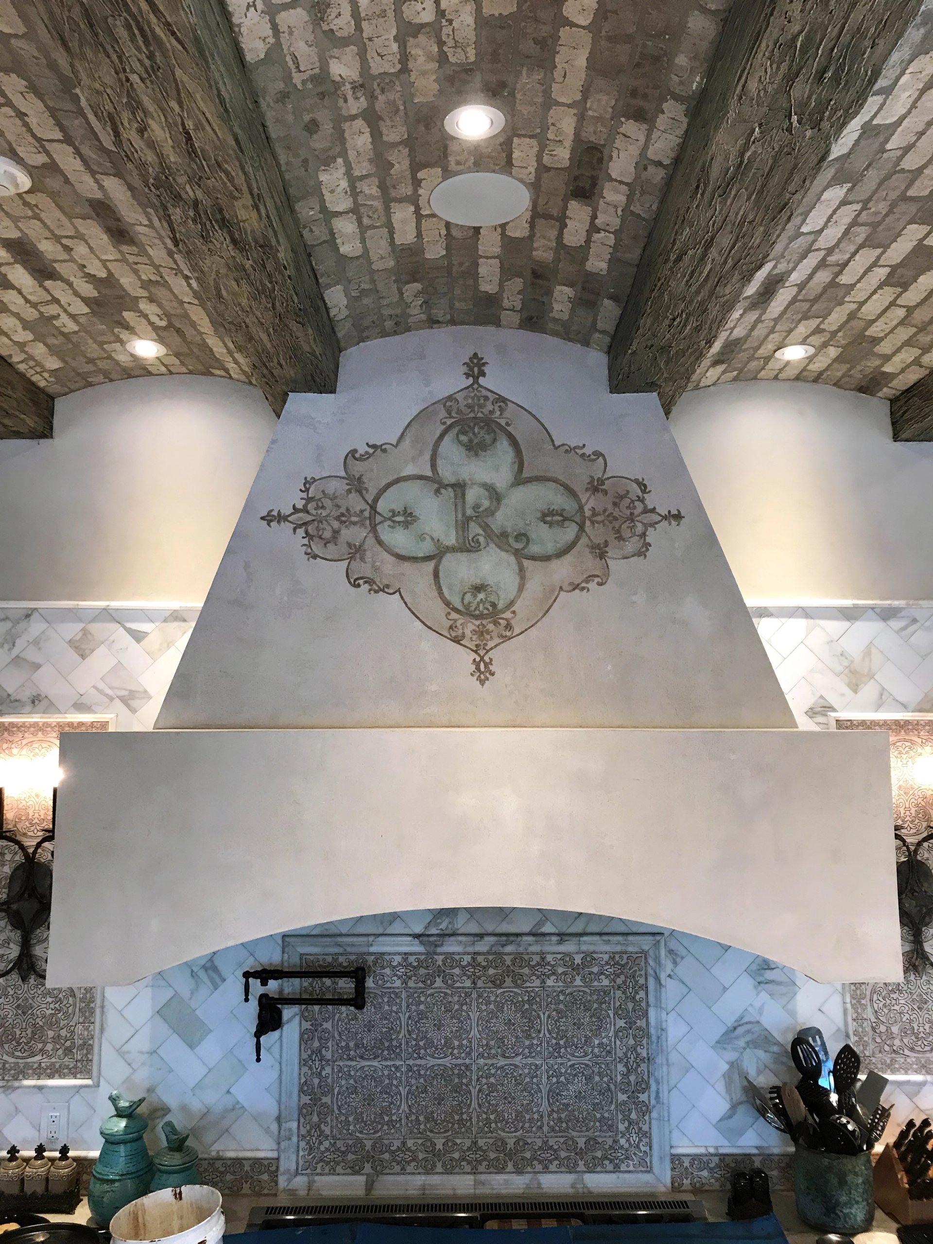 Venetian plaster range hood with hand-painted modello monogram stencil in a grand kitchen with barrel-vault brick ceiling