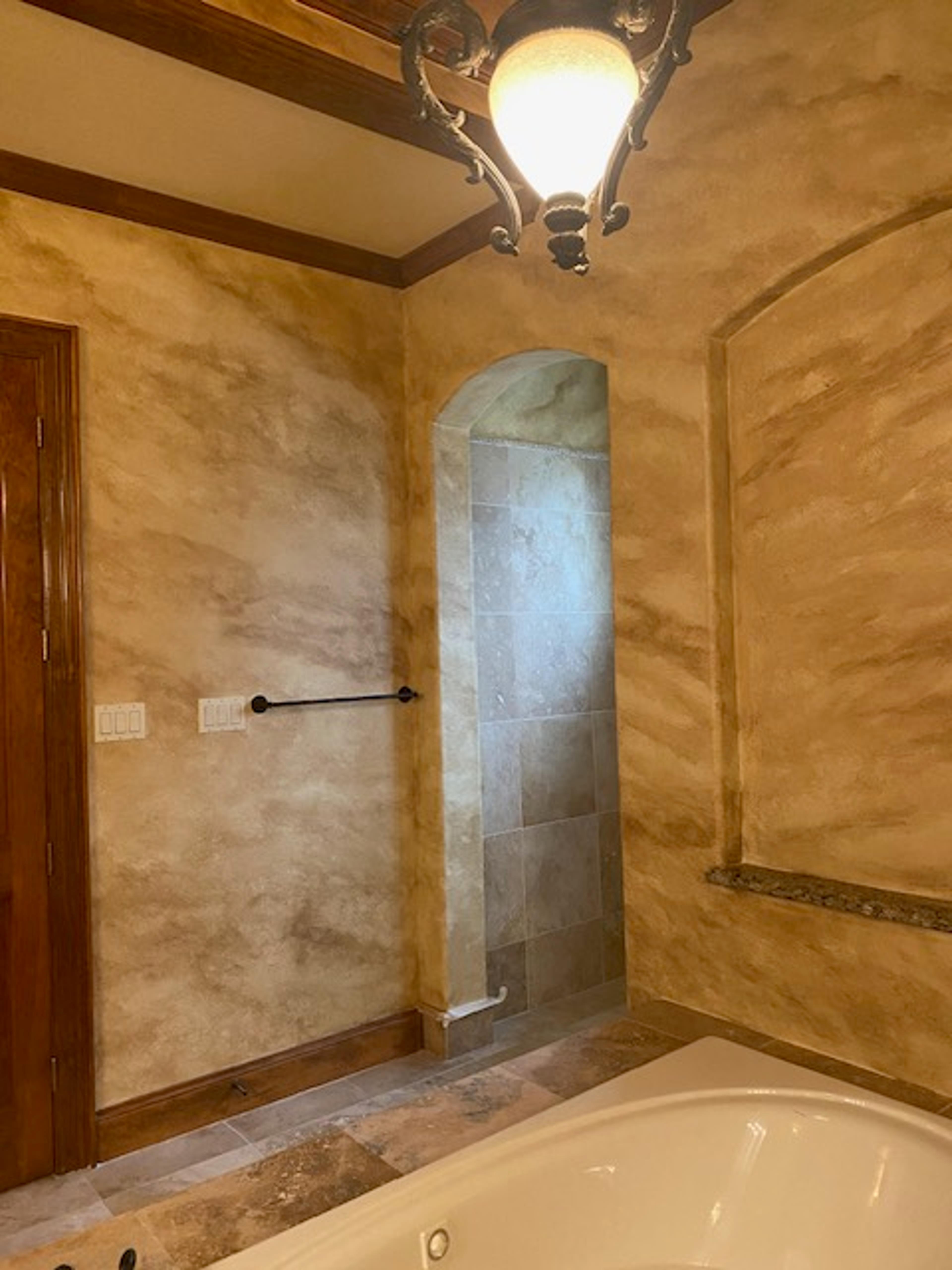 Aged travertine faux finish in warm amber tones covering full bathroom walls with arched tile niche and soaking tub, Houston TX