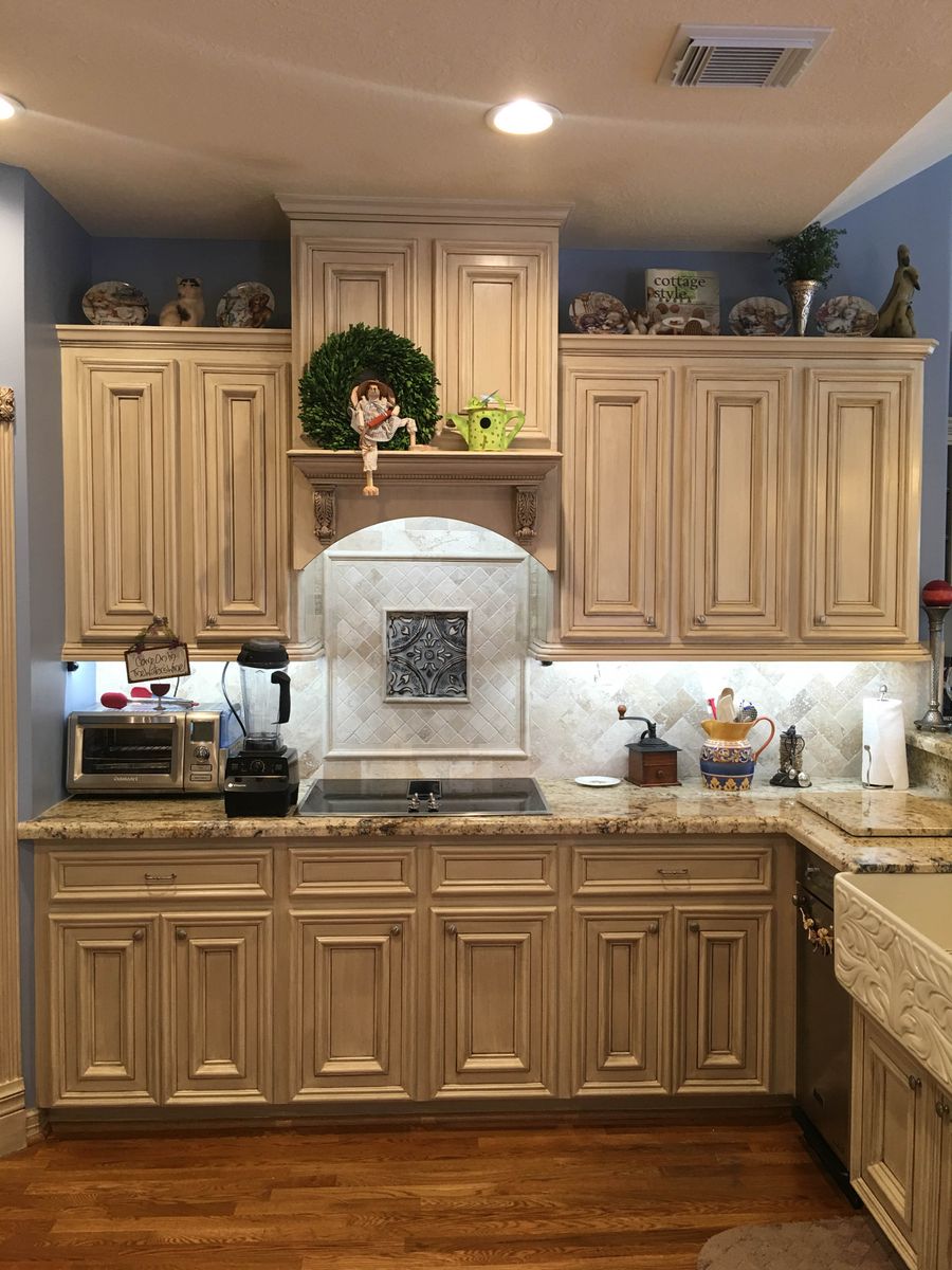 Antiqued cream glazed kitchen cabinetry with carved range hood corbels, marble herringbone backsplash, and embossed tin medallion, Houston.